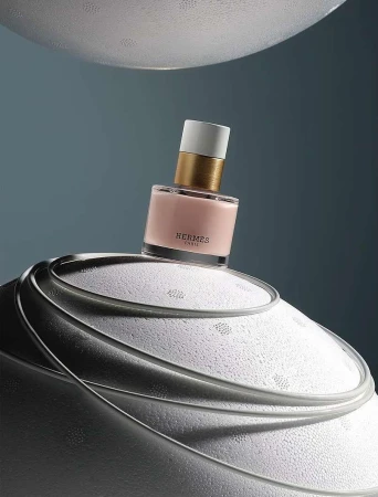 Hermes perfume bottle on a textured white surface against a blue backdrop. The bottle features a white cap, gold neck, and light pink liquid.