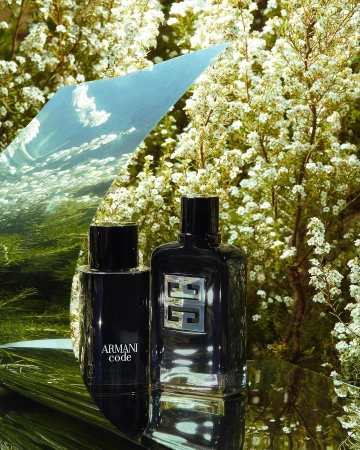 Two perfume bottles placed on a reflective surface outdoors against a backdrop of small white flowers and foliage.