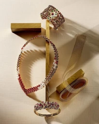 Fine jewelry arrangement featuring ruby and pearl necklace, jeweled cuffs, and gold mesh ribbon on textured paper backdrop.