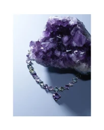 Multigemstone link necklace resting below a large raw purple amethyst crystal on a pale grey studio background.