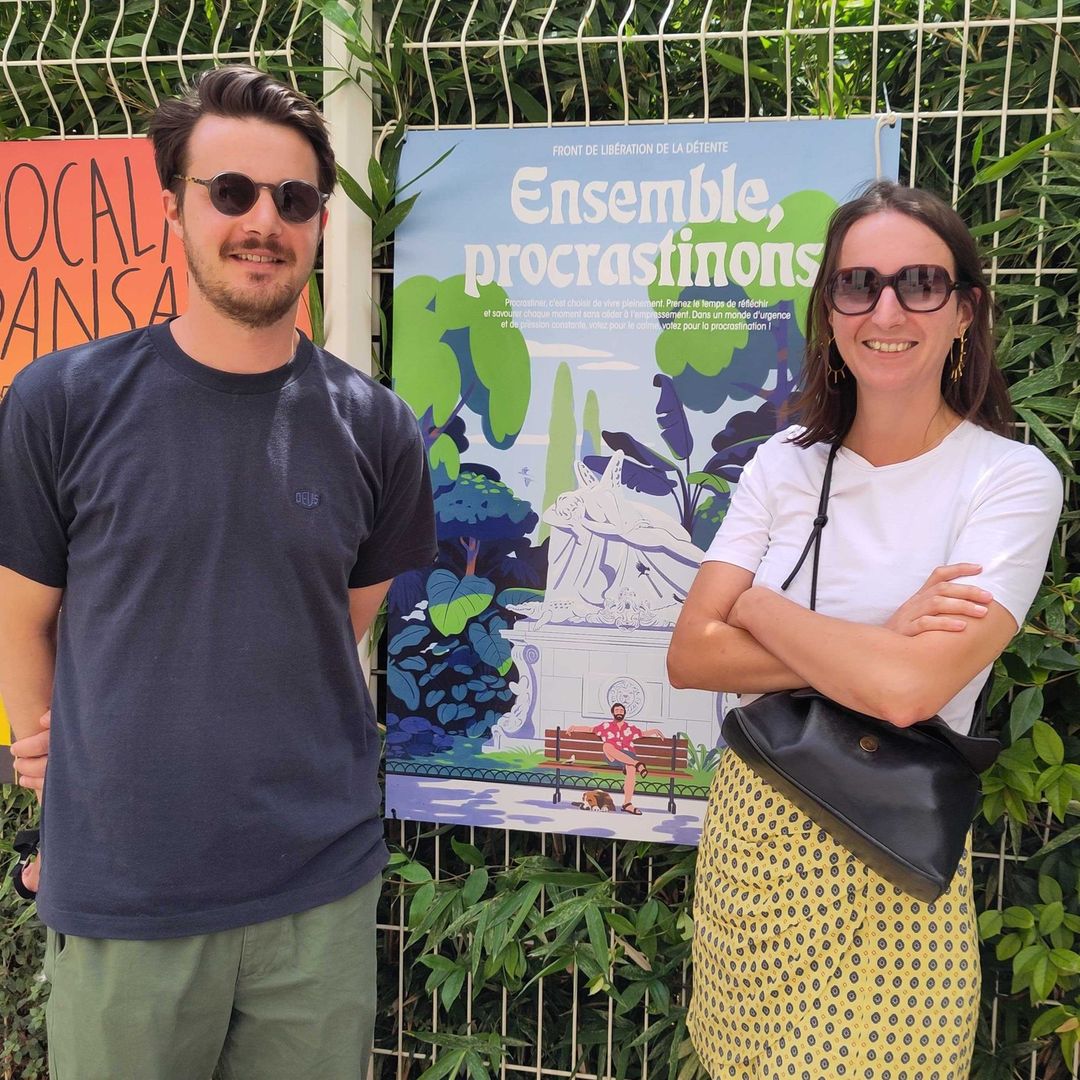Two smiling adults stand in front of an illustrated poster in an outdoor setting with natural daylight. The poster features a colorful design.