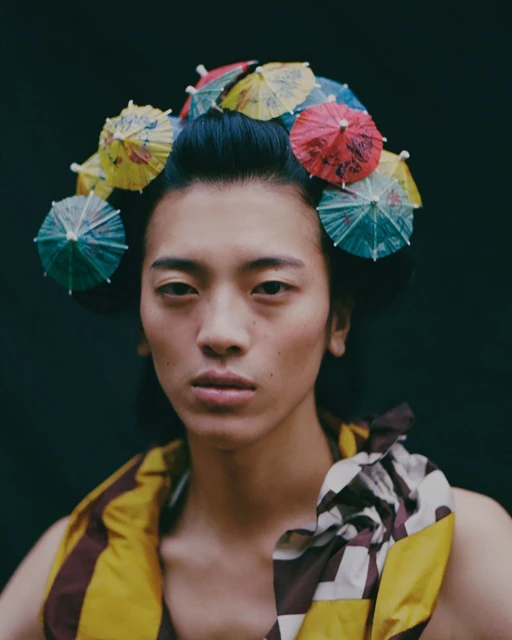 Portrait of an Asian adult with colorful cocktail umbrellas in their hair, wearing a brown and yellow garment against a dark backdrop.