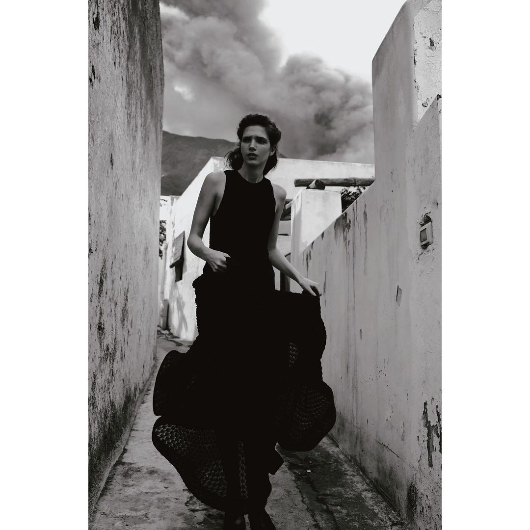 Monochrome fashion shot of an adult woman wearing a flowing black dress, posing in a narrow alley with a smoky background.