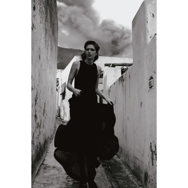 Monochrome fashion shot of an adult woman wearing a flowing black dress, posing in a narrow alley with a smoky background.