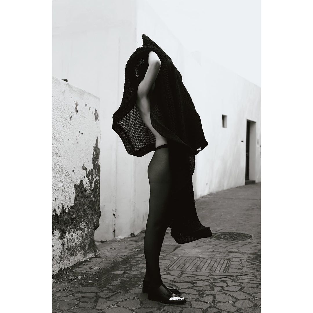 Monochrome fashion shot of a slender woman in a knitted cover-up, visible panties and stockings, walking along a tiled urban walkway.