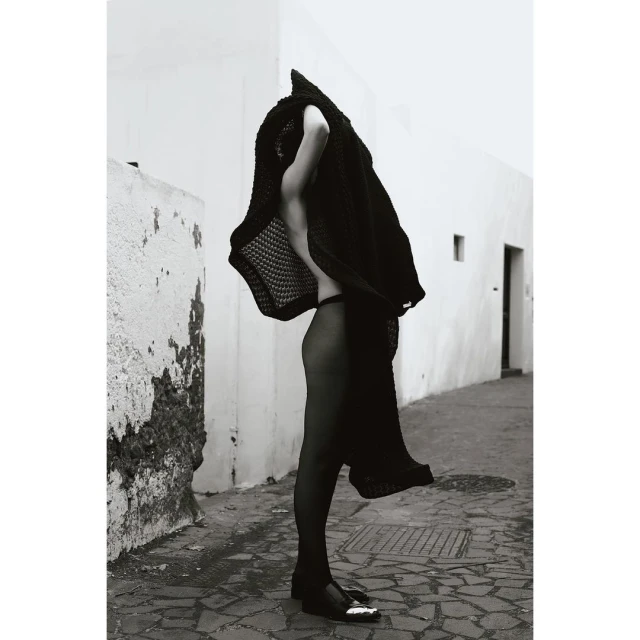 Monochrome fashion shot of a slender woman in a knitted cover-up, visible panties and stockings, walking along a tiled urban walkway.