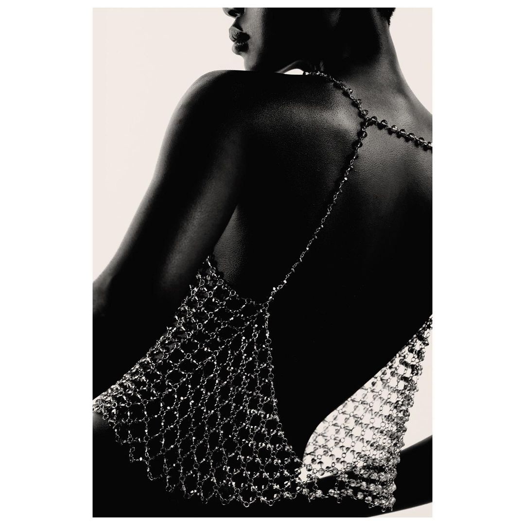 Back view of a woman wearing a metal mesh top, posing in front of a neutral backdrop, emphasizing fashion and minimalist style.