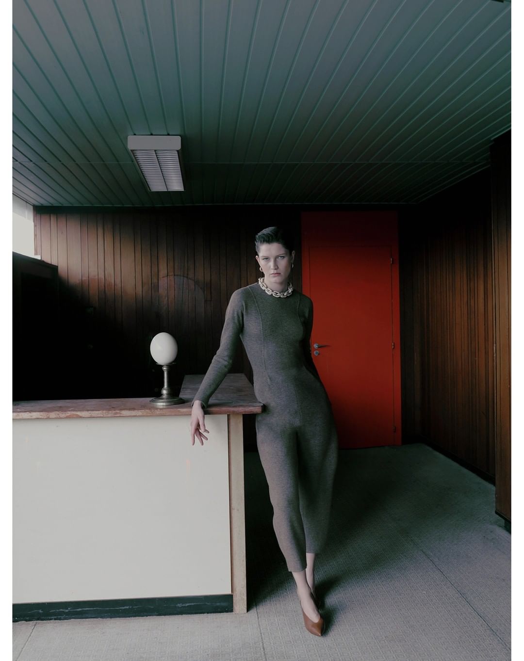 Indoor shot of an adult woman in a gray dress and brown shoes, leaning against a counter in a room with green ceiling and wood walls.