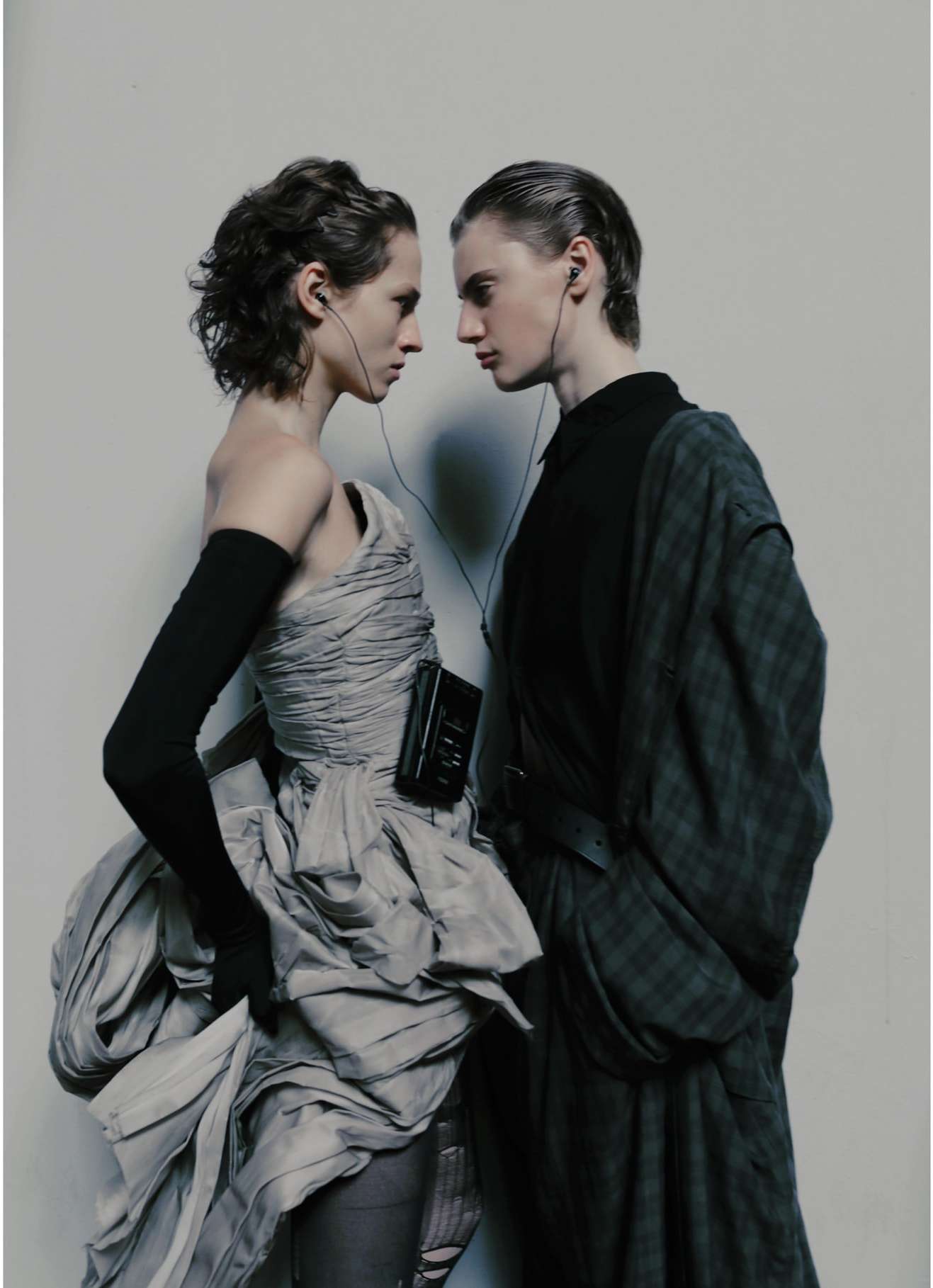 Two fashion models in a studio setting, one in an off-the-shoulder dress, the other in an oversized black and green plaid shirt.