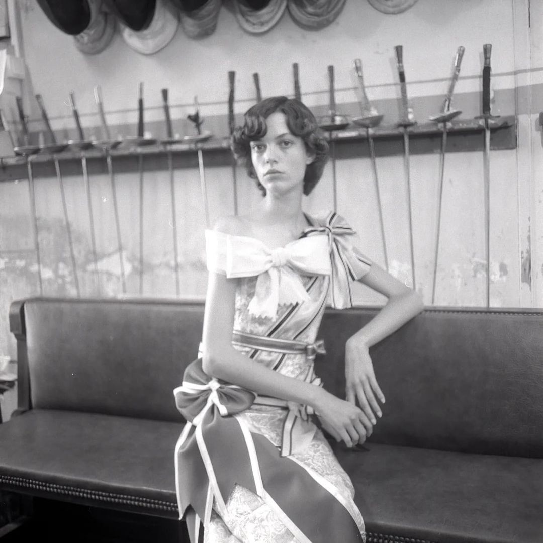 A fair-skinned adult woman with dark hair in an off-the-shoulder dress, seated on a bench against a wall with fencing equipment hanging on it.