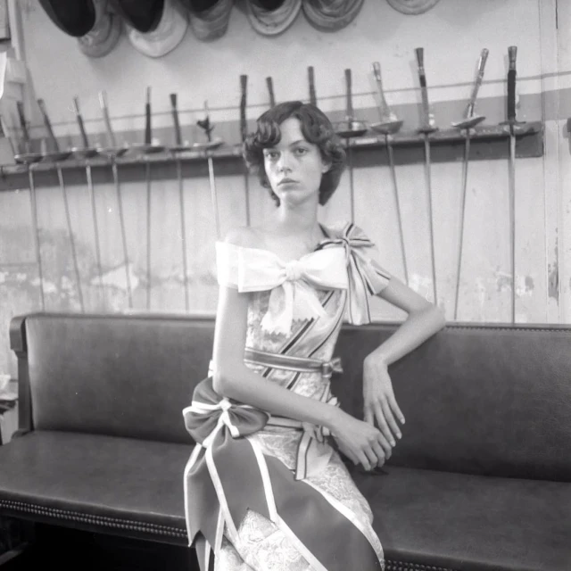 A fair-skinned adult woman with dark hair in an off-the-shoulder dress, seated on a bench against a wall with fencing equipment hanging on it.