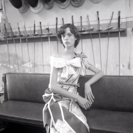 A fair-skinned adult woman with dark hair in an off-the-shoulder dress, seated on a bench against a wall with fencing equipment hanging on it.