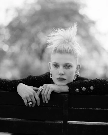 Black and white portrait of a woman with spiky blonde hair resting her chin on crossed arms over a dark slatted bench.