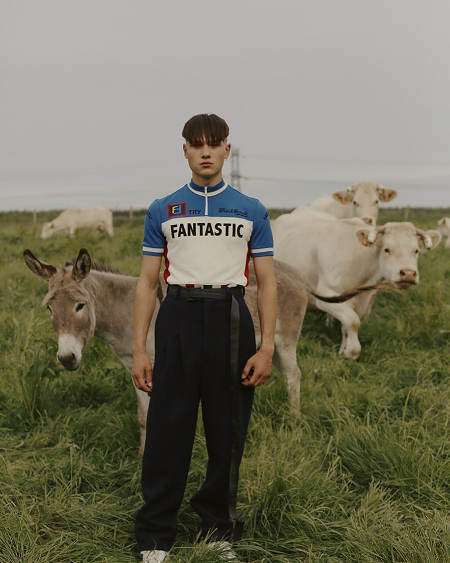 Young man poses in a field with donkeys and cows, wearing a retro-style jersey and dark trousers, capturing a pastoral, fashion-forward scene.