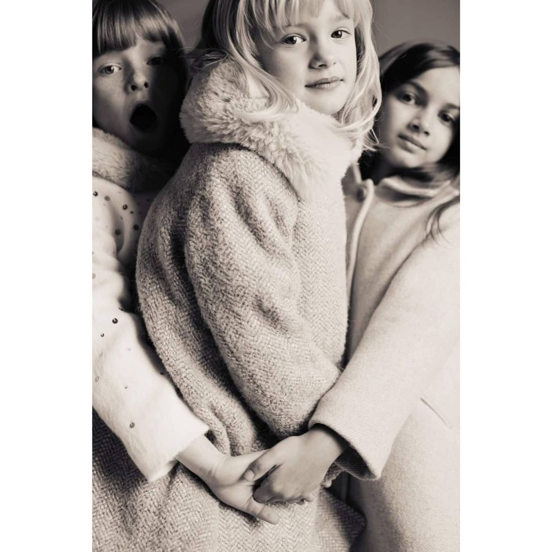 A black-and-white portrait features three young girls in winter coats, two embracing, one making an open-mouthed expression. 
