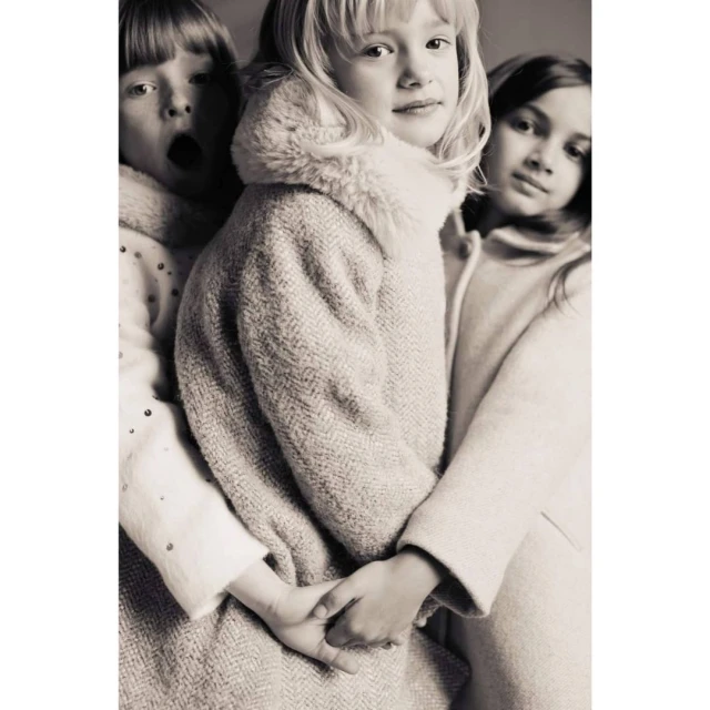 A black-and-white portrait features three young girls in winter coats, two embracing, one making an open-mouthed expression. 