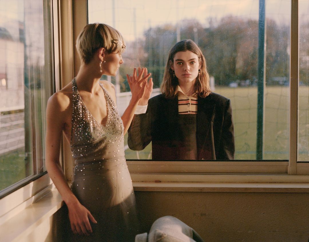Two adult women pose indoors with natural lighting. One woman wears a halter dress, and the other wears a blazer and collared shirt.