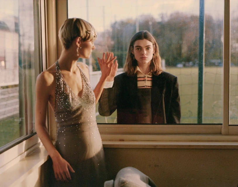 Two adult women pose indoors with natural lighting. One woman wears a halter dress, and the other wears a blazer and collared shirt.