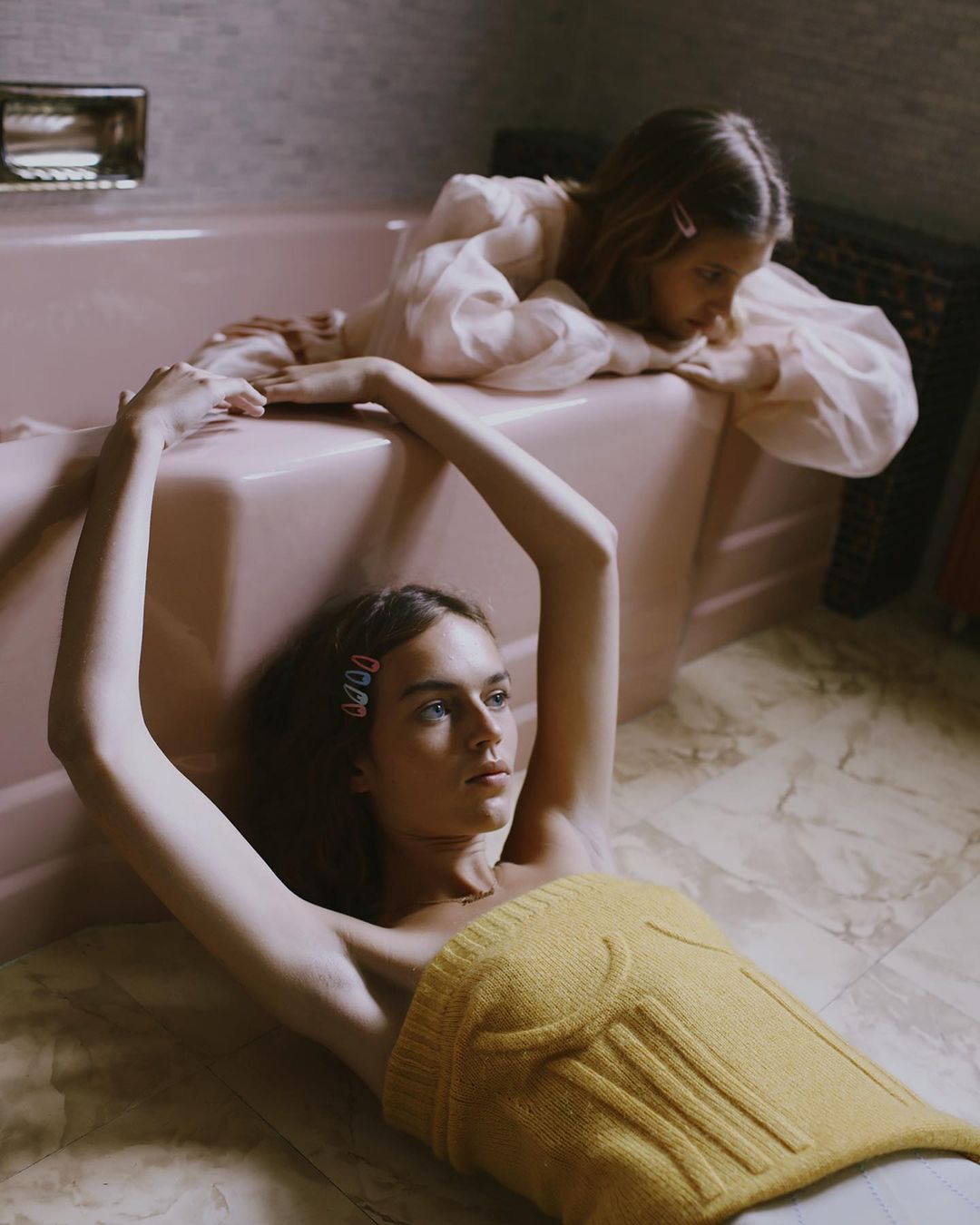 Two women in a pink bathroom: One lies on the tile floor in a yellow top, while the other reclines on the edge of a pink bathtub.