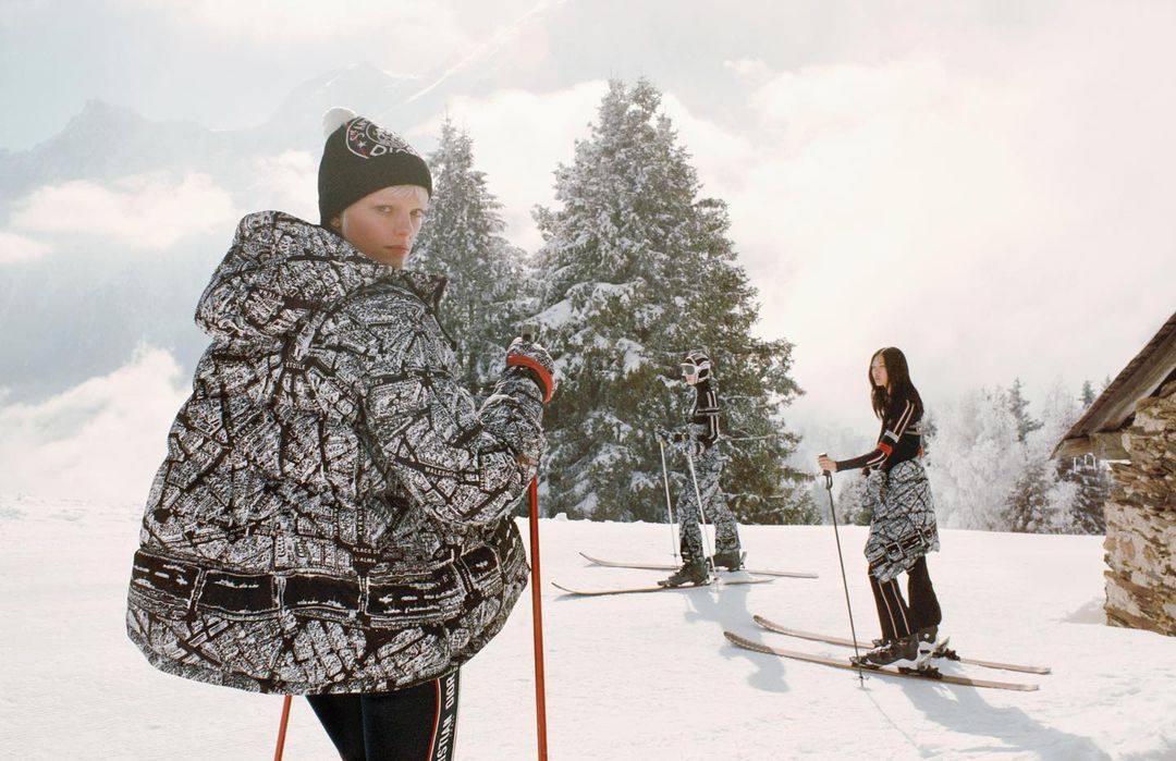 Three people in patterned outerwear and hats, skiing on a snowy mountain with pine trees and clouds in the background on a sunny day.