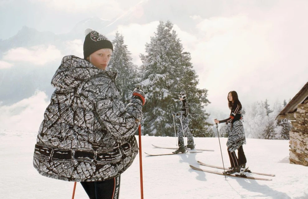 Three people in patterned outerwear and hats, skiing on a snowy mountain with pine trees and clouds in the background on a sunny day.