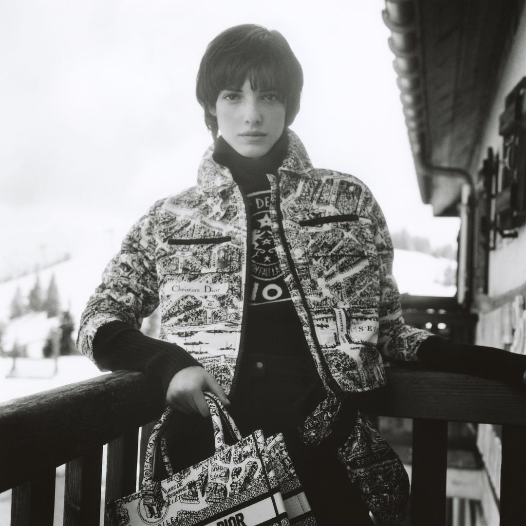 Woman in black-and-white printed jacket and black turtleneck holds a Dior tote bag on a balcony with snowy mountains visible.