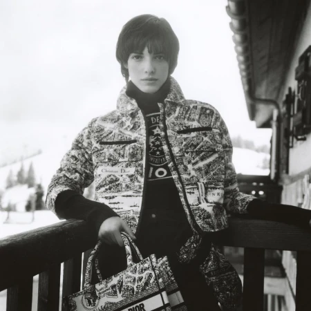 Woman in black-and-white printed jacket and black turtleneck holds a Dior tote bag on a balcony with snowy mountains visible.