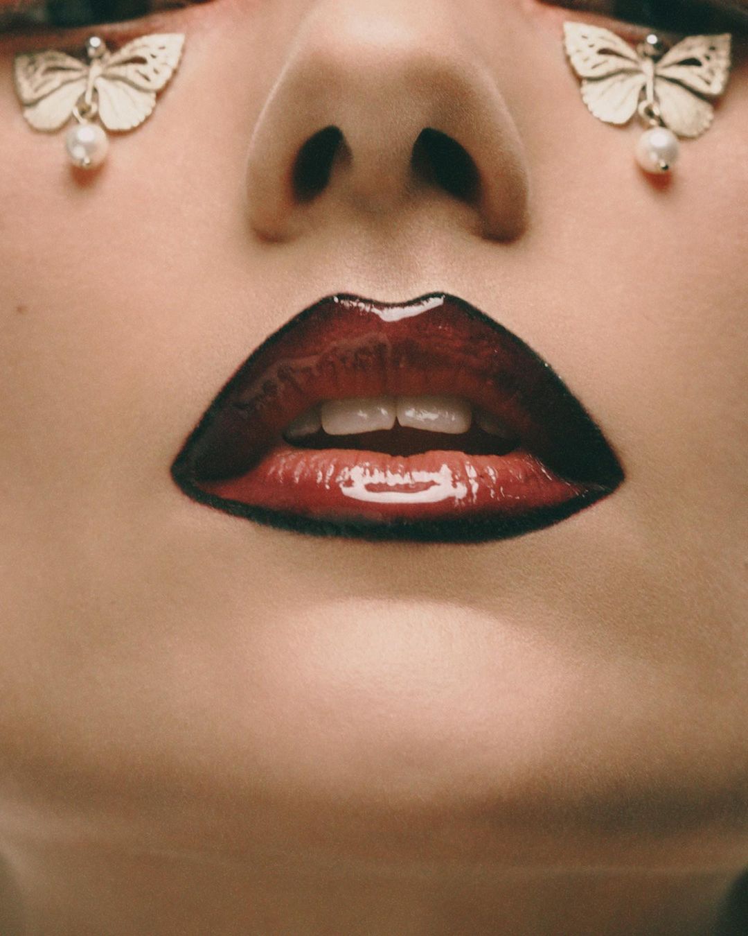 Close-up of a woman's face featuring glossy red lips and butterfly-shaped earrings with pearl drops. Beauty shot.