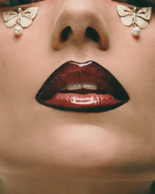 Close-up of a woman's face featuring glossy red lips and butterfly-shaped earrings with pearl drops. Beauty shot.