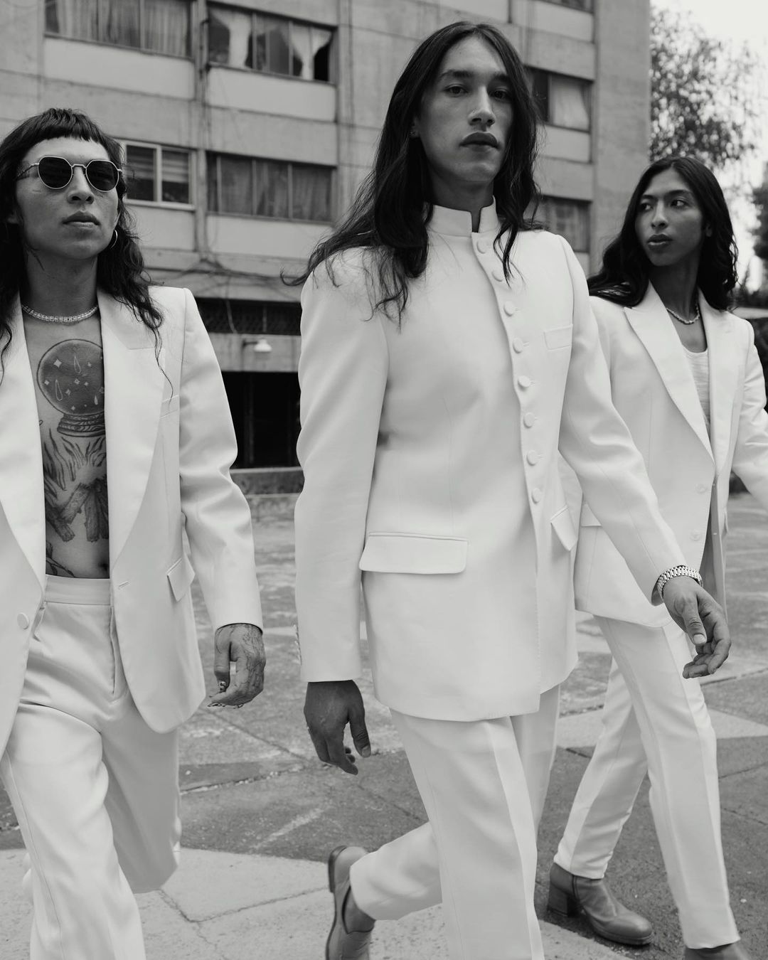 Three fashionably dressed men in white suits walk together in an urban setting. The monochrome image highlights their long hair and outfits.