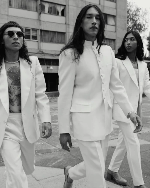 Three fashionably dressed men in white suits walk together in an urban setting. The monochrome image highlights their long hair and outfits.