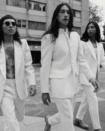 Three fashionably dressed men in white suits walk together in an urban setting. The monochrome image highlights their long hair and outfits.