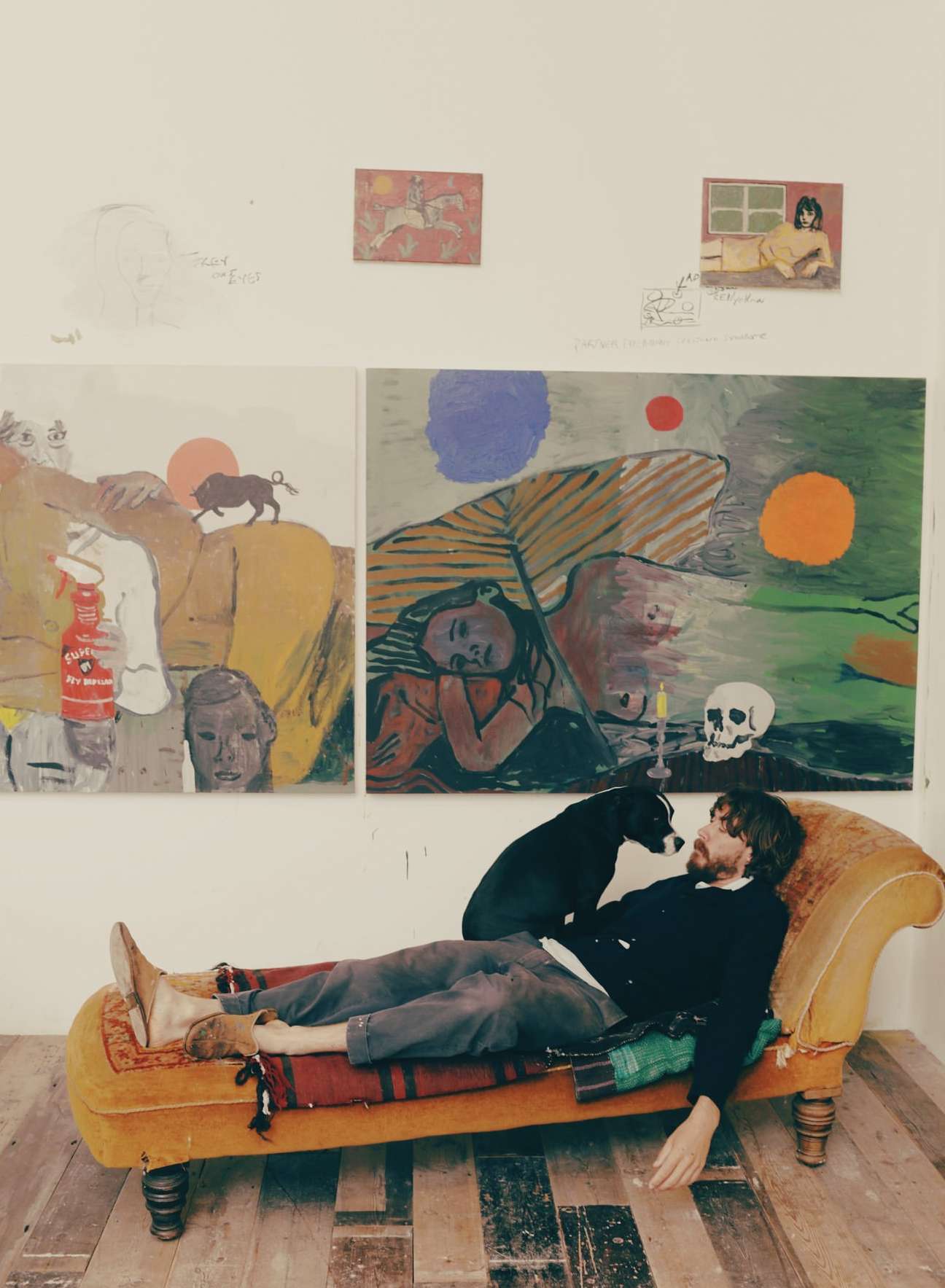 Man lounges on a chaise lounge with a dog indoors, surrounded by paintings on the wall and hardwood floors, in a brightly lit space.