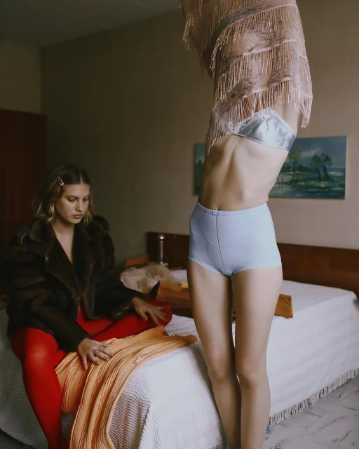 Two women pose in an indoor setting with muted lighting; one sits on a bed wearing a dark jacket and red tights, the other stands.