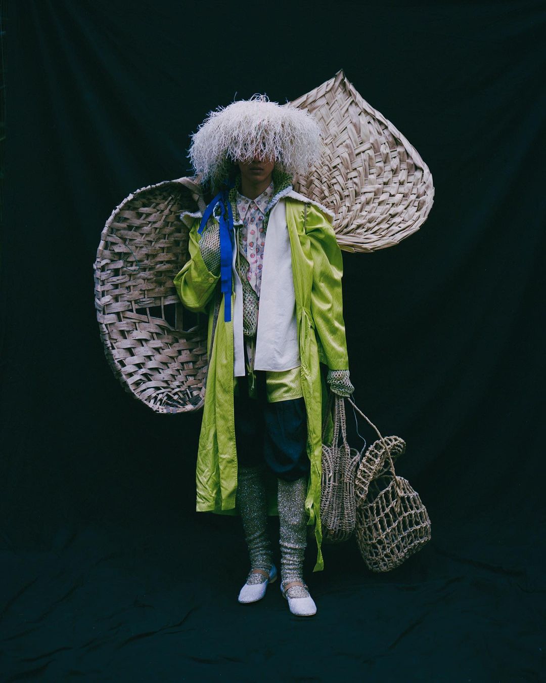 Person in a light green coat, textured stockings, and a feathery hat, holding bags against a dark background with wicker decorations.