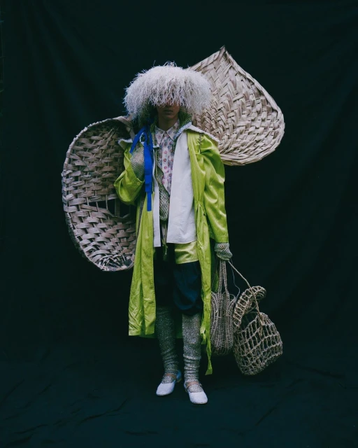 Person in a light green coat, textured stockings, and a feathery hat, holding bags against a dark background with wicker decorations.