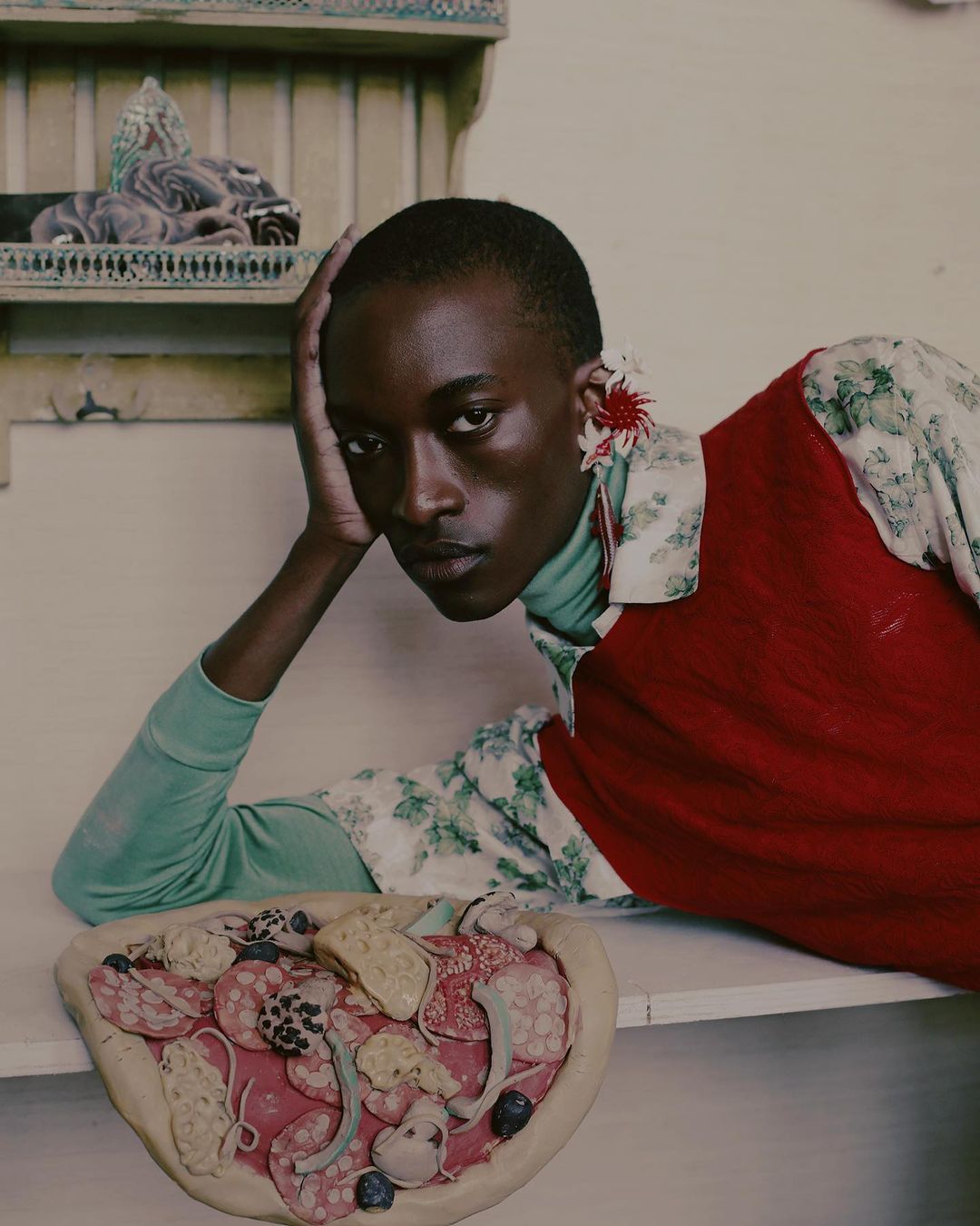 Adult fashion model wearing floral print shirt and red top poses with pizza on a light surface indoors with shelf and display behind.
