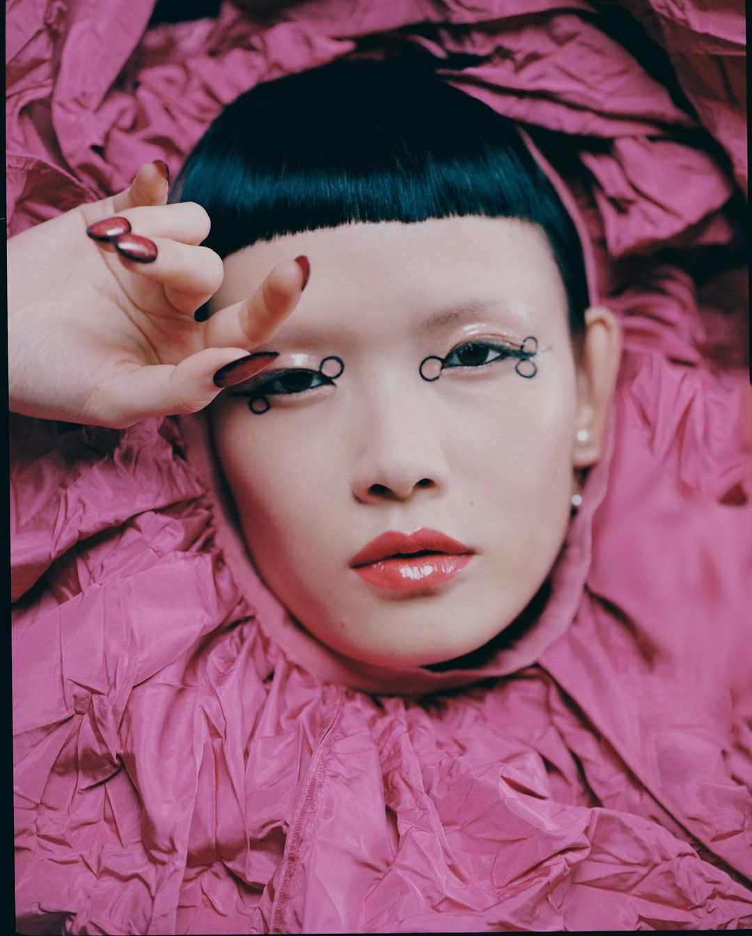 Close-up portrait of a woman with geometric eyeliner and bold lipstick, wearing a voluminous pink garment, against a matching background.