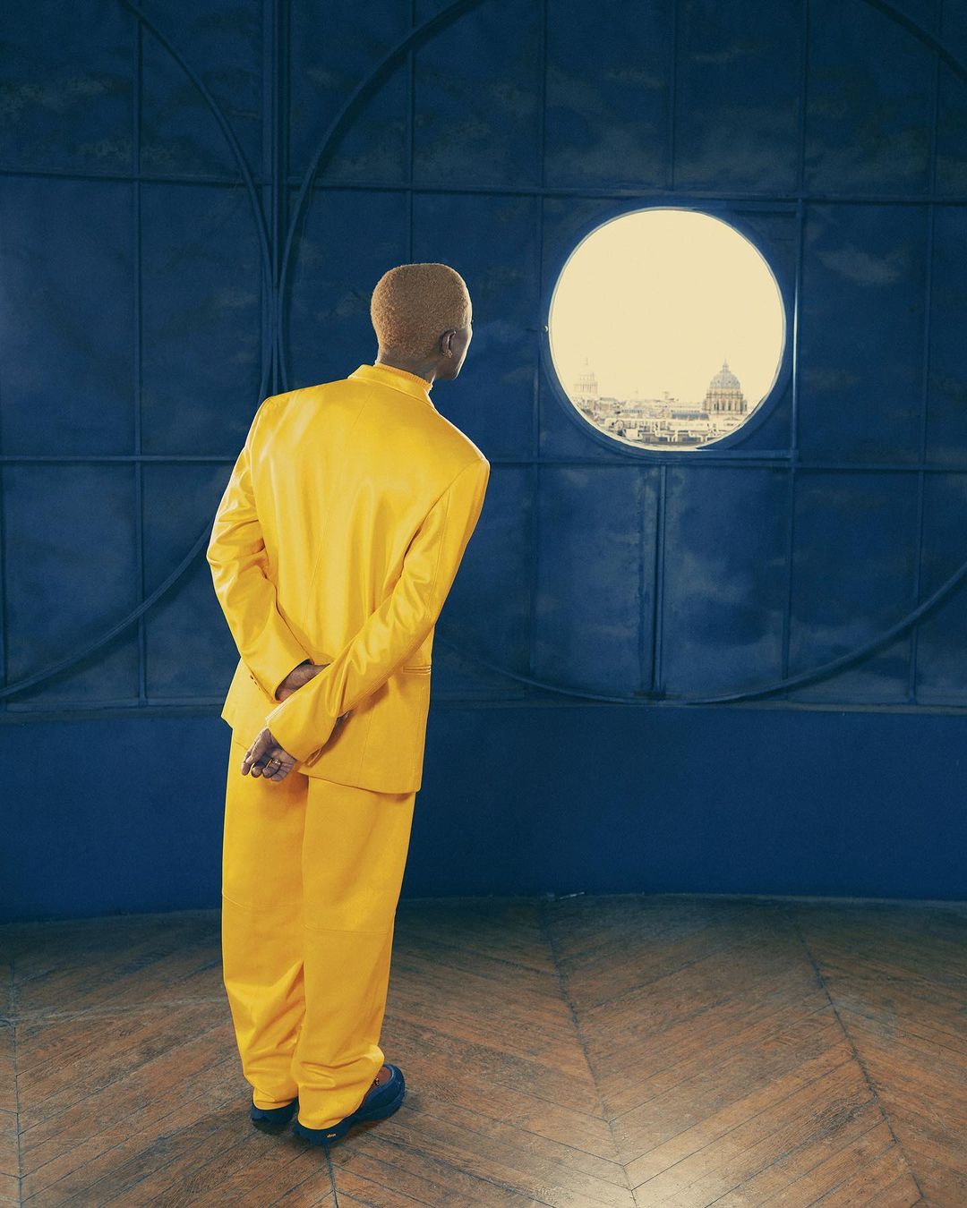 Blond adult in yellow suit stands in room, back to camera, looking out round window at cityscape under bright daylight.