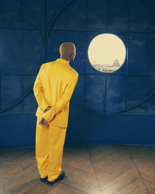 Blond adult in yellow suit stands in room, back to camera, looking out round window at cityscape under bright daylight.