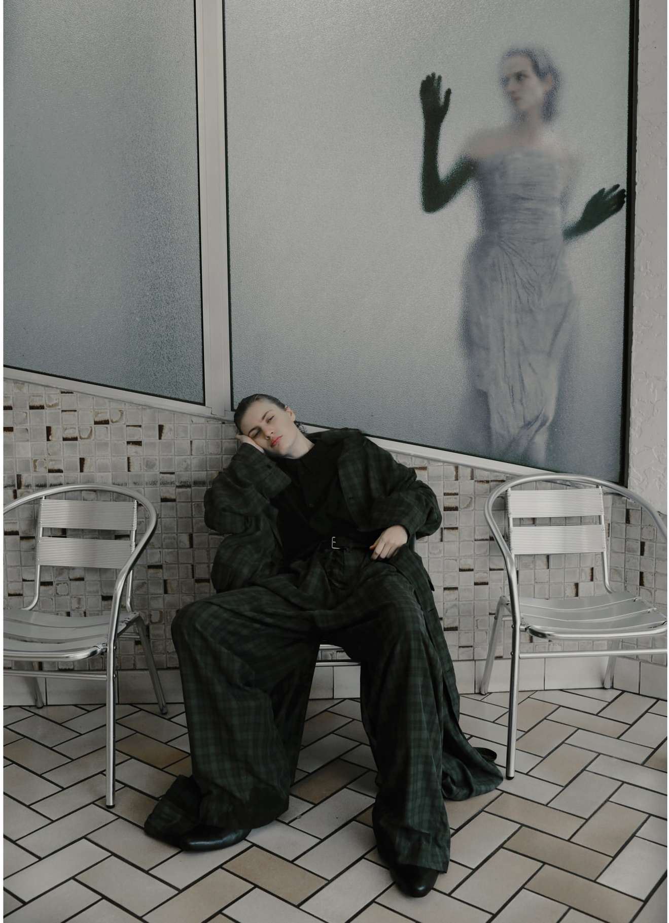 Indoor fashion shot of a person in a dark top and green plaid pants sitting on a metal chair, with blurred figure in background.
