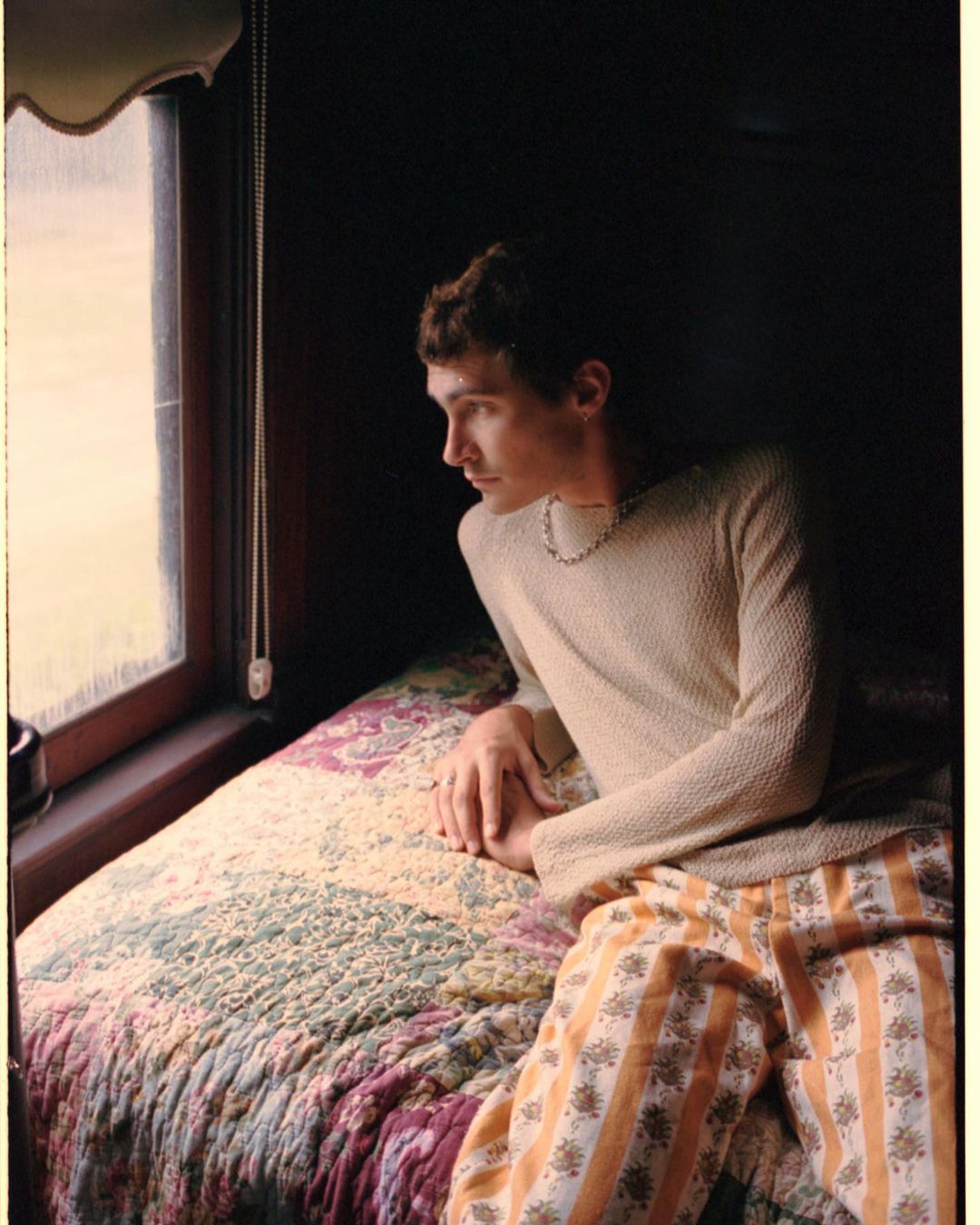 Young man looks out window from bed in photo studio with indoor, soft lighting. He is wearing a knit top and striped floral print pants.