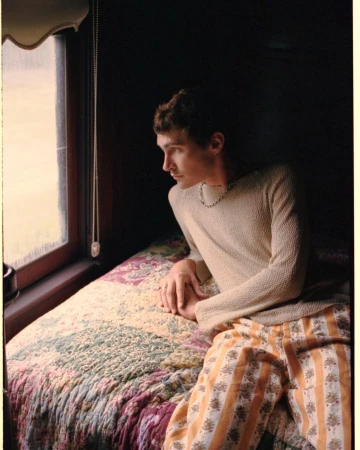 Young man looks out window from bed in photo studio with indoor, soft lighting. He is wearing a knit top and striped floral print pants.