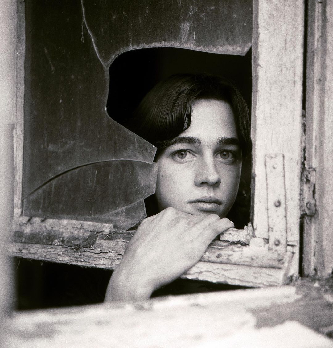 Monochrome portrait of a young adult gazing through a shattered window, creating a stark contrast between vulnerability and decay.
