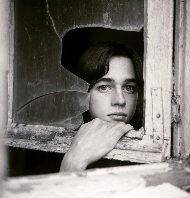 Monochrome portrait of a young adult gazing through a shattered window, creating a stark contrast between vulnerability and decay.