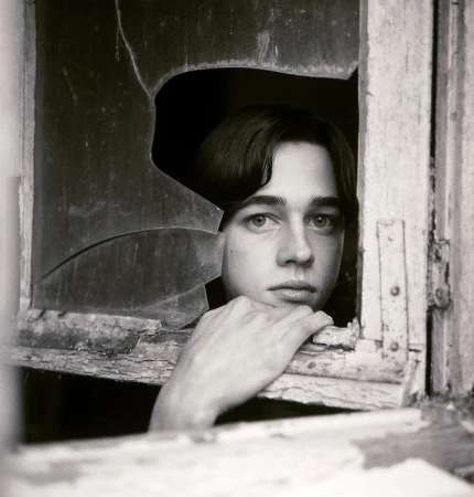 Monochrome portrait of a young adult gazing through a shattered window, creating a stark contrast between vulnerability and decay.