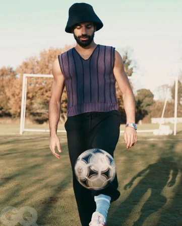 Man in sleeveless top and bucket hat juggling a soccer ball on a grassy field with soccer goal in the background under sky.