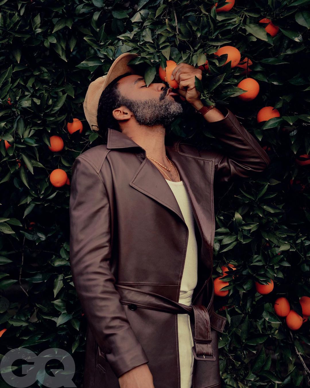 Man in a brown trench coat stands in front of an orange tree with fruit, eyes closed, as he tastes an orange.
