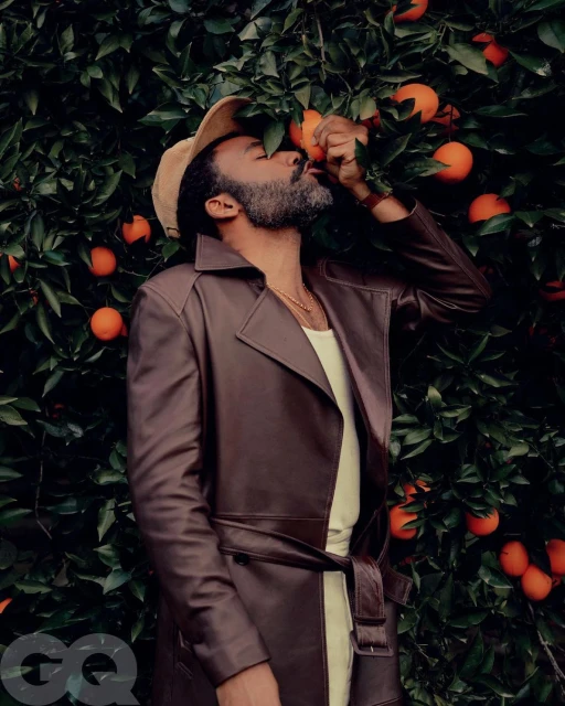 Man in a brown trench coat stands in front of an orange tree with fruit, eyes closed, as he tastes an orange.