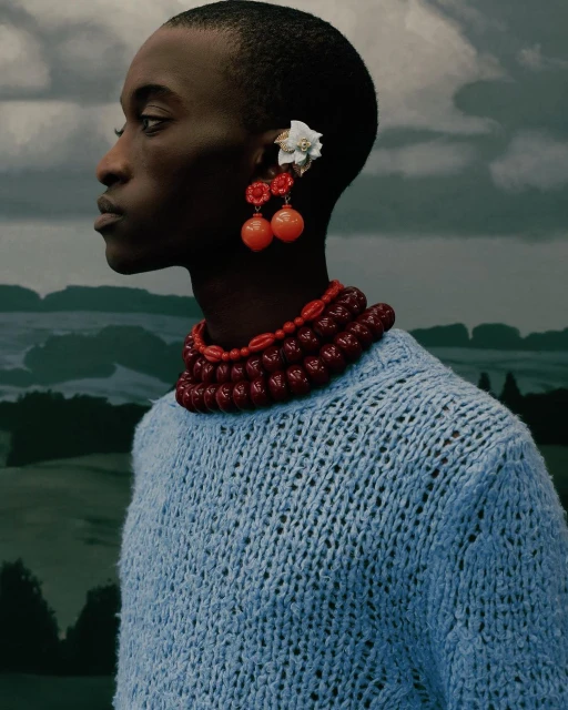 Portrait of a Black adult woman wearing a blue sweater and red beaded jewelry, posed in front of a landscape backdrop.